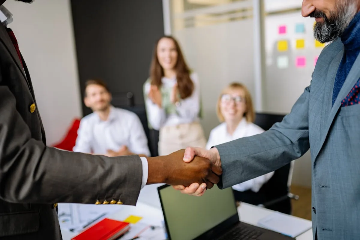 Two professionals shaking hands as colleagues applaud in the background